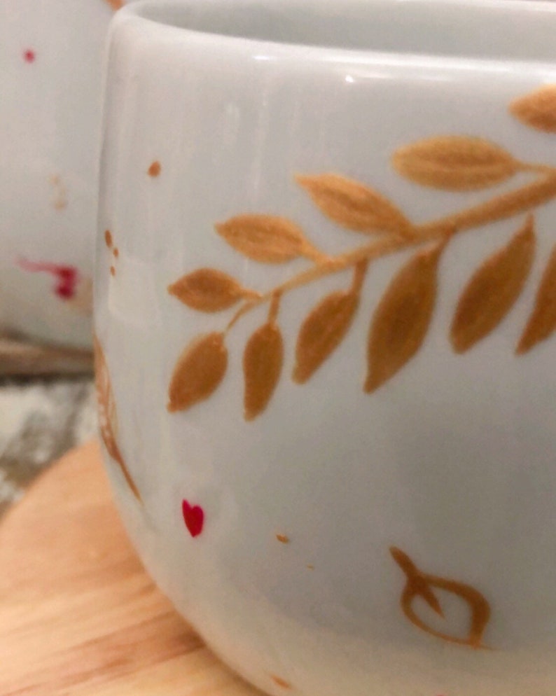 May include: Close-up of a white ceramic mug with a gold leaf design. The mug has a rounded shape and is decorated with gold-colored leaves and small red heart accents. The mug is on a wooden surface.