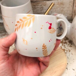 May include: White ceramic mug with a gold-colored leaf design and red speckles. A gold-colored spoon rests inside the mug. The mug is held in a hand, with a wooden coaster beneath it. Other mugs are visible in the background.