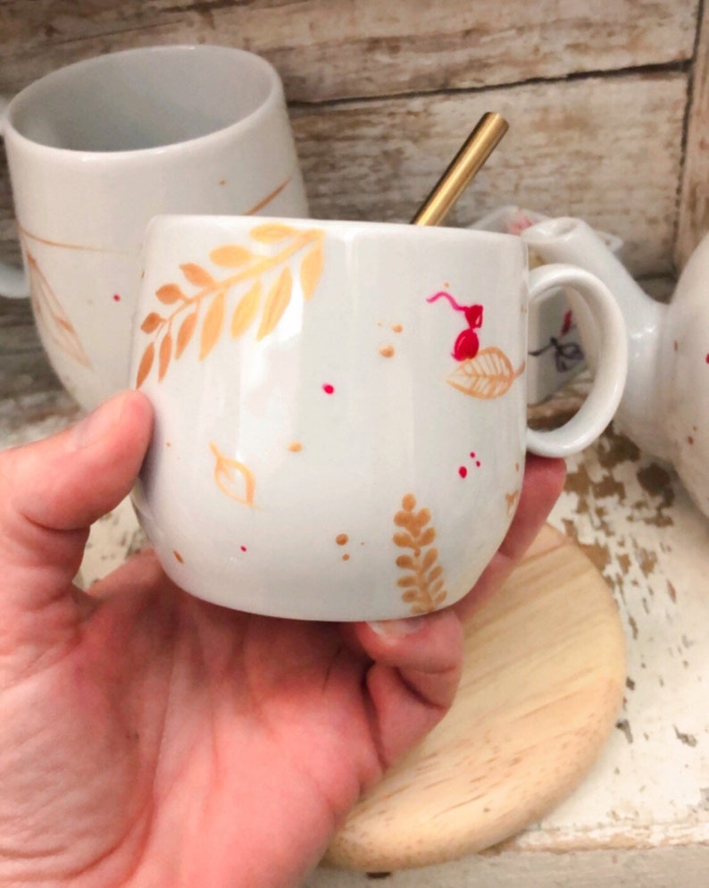 May include: White ceramic mug with a gold-colored leaf design and red speckles. A gold-colored spoon rests inside the mug. The mug is held in a hand, with a wooden coaster beneath it. Other mugs are visible in the background.