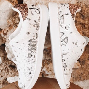 May include: A pair of white canvas trainers with black floral designs and gold glitter accents. The left shoe has the word "Bride" written on it.