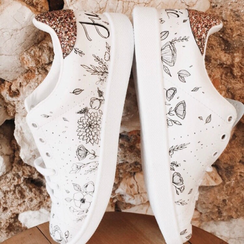 May include: A pair of white canvas trainers with black floral designs and gold glitter accents. The left shoe has the word "Bride" written on it.