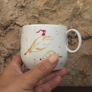 May include: A white ceramic mug with a rounded shape and a handle. The mug is decorated with brown leaf designs and red speckles. The mug is held by a person's hand.