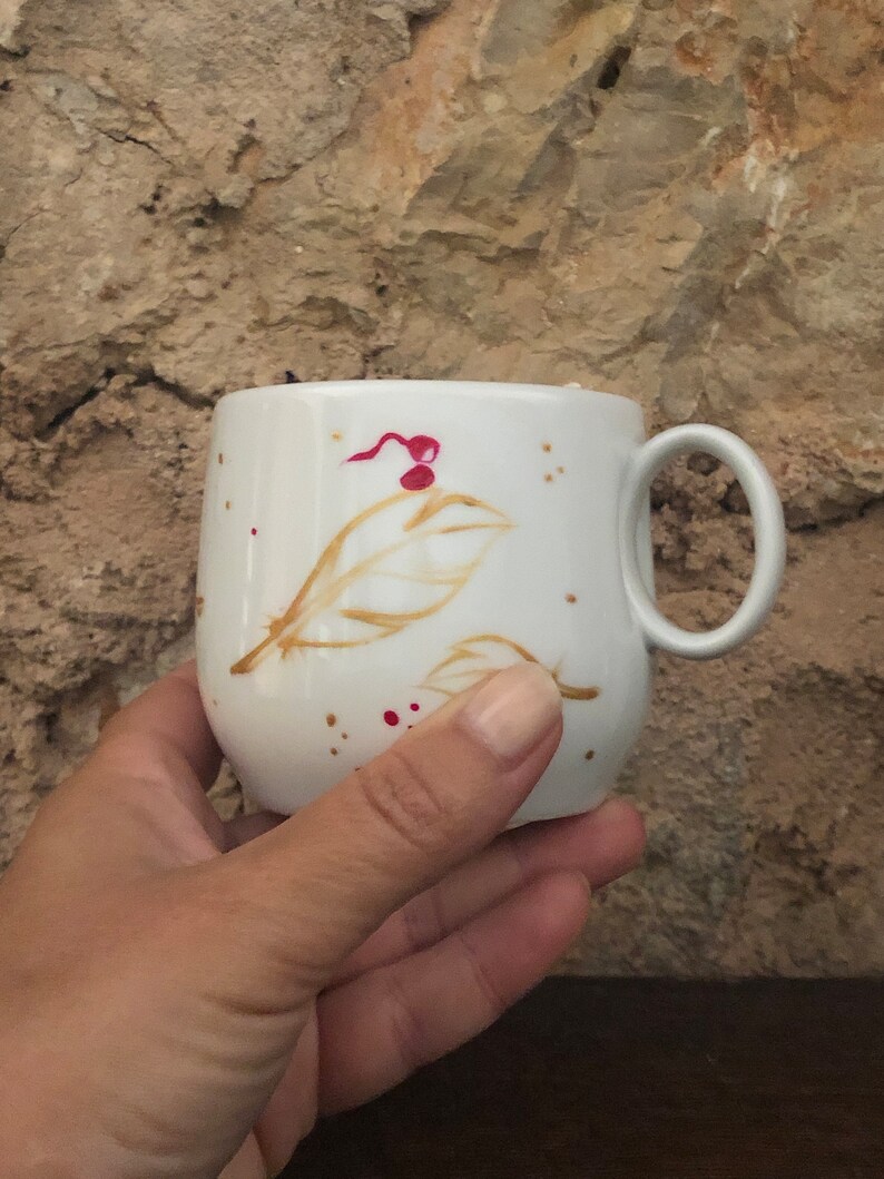 May include: A white ceramic mug with a rounded shape and a handle. The mug is decorated with brown leaf designs and red speckles. The mug is held by a person's hand.