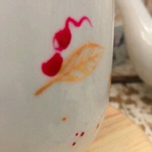 May include: White ceramic mug with a floral design. The mug features a red and orange leaf and flower pattern. Small red dots are scattered around the design. The mug is sitting on a wooden surface.