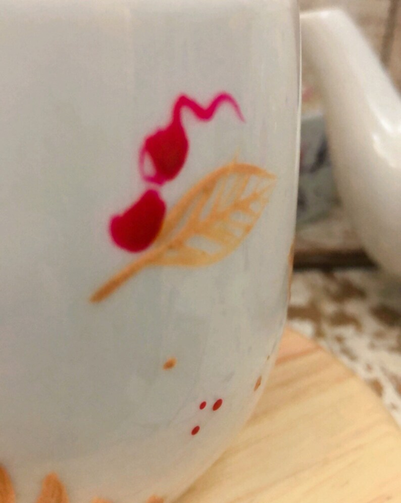 May include: White ceramic mug with a floral design. The mug features a red and orange leaf and flower pattern. Small red dots are scattered around the design. The mug is sitting on a wooden surface.