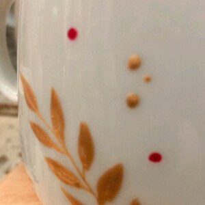 May include: White ceramic mug with a decorative design. The mug features a branch of orange leaves and small orange and red dots. The handle is visible on the left side of the mug.