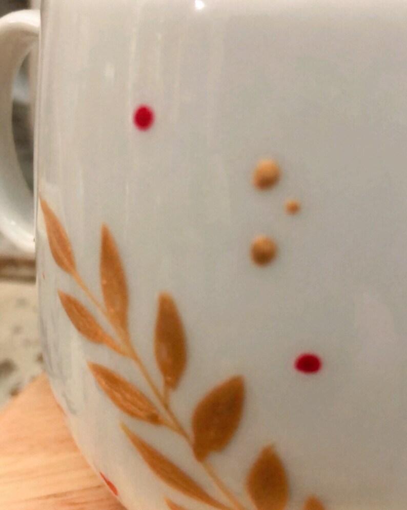 May include: White ceramic mug with a decorative design. The mug features a branch of orange leaves and small orange and red dots. The handle is visible on the left side of the mug.