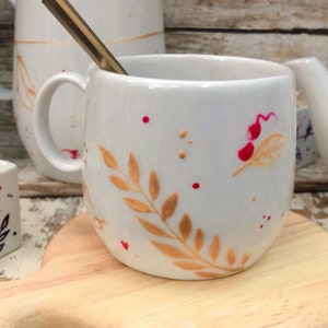 May include: White ceramic mug with a gold-colored spoon inside. The mug features a hand-painted design of gold leaves and red dots. The mug is sitting on a wooden cutting board. Other mugs are in the background.
