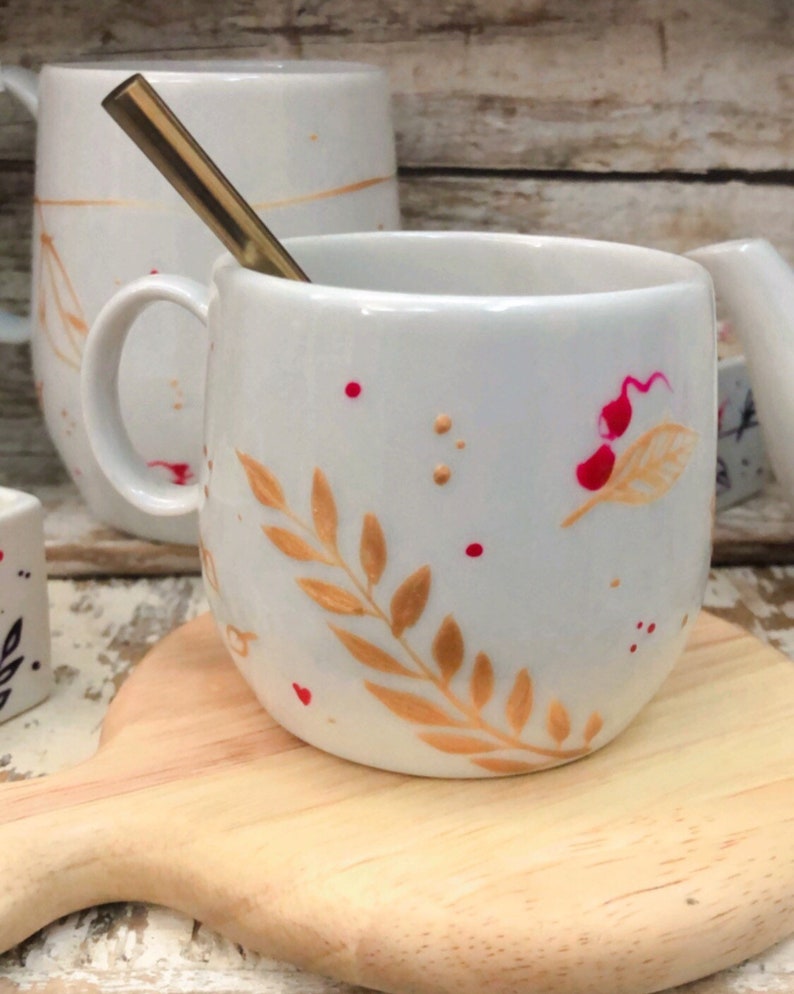 May include: White ceramic mug with a gold-colored spoon inside. The mug features a hand-painted design of gold leaves and red dots. The mug is sitting on a wooden cutting board. Other mugs are in the background.