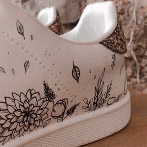 May include: White canvas trainers with a black floral design and gold glitter accents on the heel.
