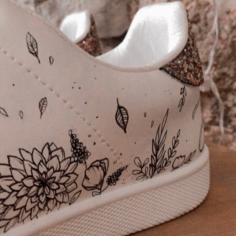 May include: White canvas trainers with a black floral design and gold glitter accents on the heel.