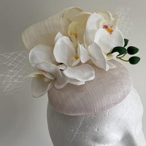 May include: A cream-coloured fascinator adorned with white orchid flowers with yellow and pink details. The hat has a pale pink base and a cream-coloured, textured top, finished with a fine veil.