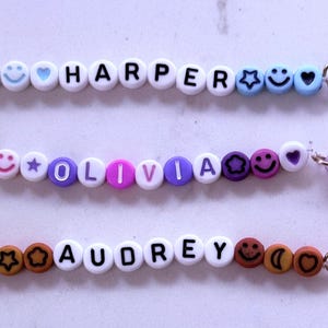 May include: Three personalised name bracelets with colourful beads and charms. The bracelets feature the names "HARPER", "OLIVIA", and "AUDREY" with smiley faces, hearts, stars and moons. Each bracelet has a silver clasp and a tassel.