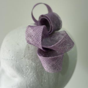 May include: A lavender headpiece with a looped design. The headpiece is made of a woven material and is attached to a white head form. The headpiece has a textured appearance, suitable for special occasions.