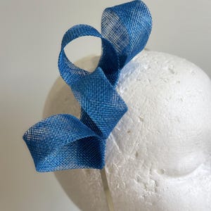 May include: A blue headband with a woven fabric design. The headband features a decorative loop and swirl design attached to a thin, silver-coloured band. The accessory is displayed on a white mannequin head.