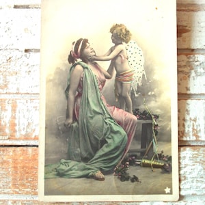 May include: Vintage postcard featuring a woman in a pink and green gown being playfully touched by a cherub with polka-dot wings. The cherub wears a striped wrap. The postcard has a faded, antique aesthetic.