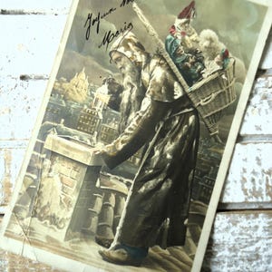 Antica cartolina di Natale con pieghe, camino sul tetto di Babbo Natale, giocattoli, bambole, lanterna serale, colori francesi del 1900