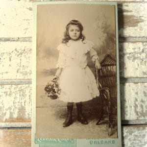May include: A sepia-toned vintage photograph of a young girl in a white dress, holding a basket of flowers and leaning on a wicker chair. The photograph is framed and has the word "ORLEANS" printed at the bottom.