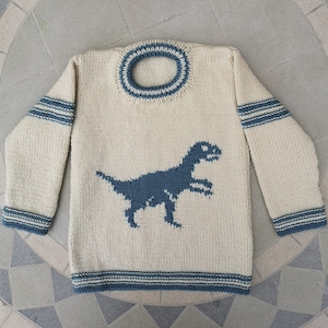 Puede incluir: Jersey de punto color crema con un diseño de dinosaurio azul. El jersey tiene rayas azules en las mangas, el cuello y el dobladillo. El dinosaurio es una silueta de un Tyrannosaurus Rex.