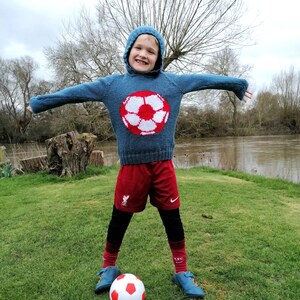 May include: A young boy wearing a blue knitted jumper with a white and red football design, red shorts, and black leggings. He is standing on a grassy field with a football at his feet.