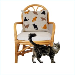 May include: A woven pillow with cat silhouettes in various colors rests on a wicker chair with a white cushion. A tabby cat with a fluffy tail stands nearby. The pillow features brown, black, and orange cat designs.