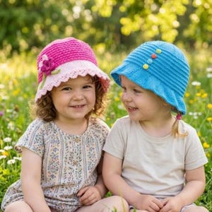 May include: Two young children wearing handmade crochet sun hats. One hat is pink with a flower, the other is blue with decorative buttons. They are sitting in a field of wildflowers, smiling at each other. The hats are a perfect accessory for sunny days.