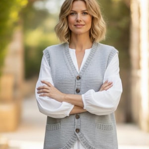 May include: A light gray knitted vest with a V-neck and four brown buttons. The vest has two front pockets and is worn over a white long-sleeved shirt. The woman is standing outside with her arms crossed.