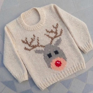 May include: A cream-colored long-sleeve knit sweater featuring a central graphic of a reindeer head. The reindeer has brown antlers, a gray face, black eyes, and a prominent red nose. The sweater has a ribbed crew neckline and cuffs.