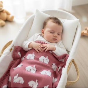May include: A sleeping baby in a white bassinet, covered with a pink blanket featuring white rabbit designs. The baby wears a white onesie and rests on a white pillow. A teddy bear is visible in the background.