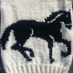 May include: A cream-colored knit garment featuring a prominent black horse silhouette pattern. The design displays a horse in a dynamic, mid-stride pose, rendered in black yarn against the light-colored, textured knitted background.