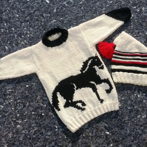 May include: A cream-colored long-sleeve sweater featuring a black horse graphic, a black ribbed collar, and black sleeve cuffs, paired with a matching cream-colored knit hat with black and red stripes and a bright red yarn pom-pom.