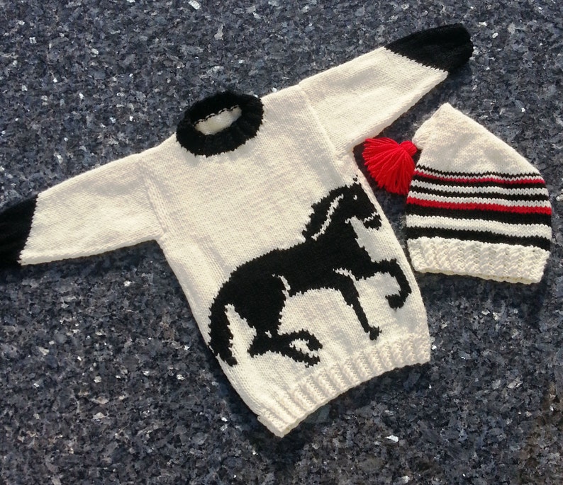 May include: A cream-colored long-sleeve sweater featuring a black horse graphic, a black ribbed collar, and black sleeve cuffs, paired with a matching cream-colored knit hat with black and red stripes and a bright red yarn pom-pom.