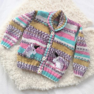 May include: A colorful, knitted cardigan with a unicorn design. The sweater features a button-up front, a collar, and two pockets, with embroidered unicorn details. The color palette includes pink, blue, purple, and gold stripes. The cardigan is displayed on a white, fluffy surface.