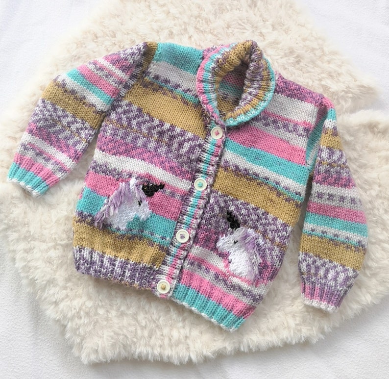May include: A colorful, knitted cardigan with a unicorn design. The sweater features a button-up front, a collar, and two pockets, with embroidered unicorn details. The color palette includes pink, blue, purple, and gold stripes. The cardigan is displayed on a white, fluffy surface.