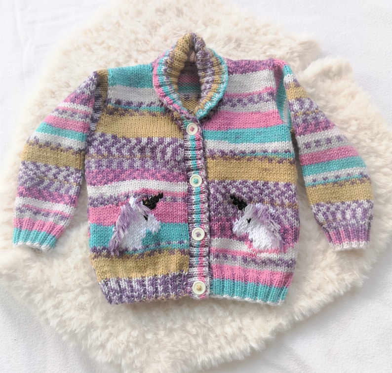 May include: A striped cardigan with a colorful pattern in shades of purple, pink, teal, and gold. The cardigan has a button closure and features two embroidered unicorn appliques on the front.