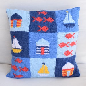 May include: A blue and white knitted cushion with a nautical theme. The cushion features images of sailboats, houses, and fish.
