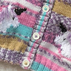 May include: Close-up of a knitted cardigan with a unicorn design. The sweater features a striped pattern in shades of pink, purple, blue, and tan. Two white unicorns with purple manes are embroidered on the front. The cardigan has three white buttons.