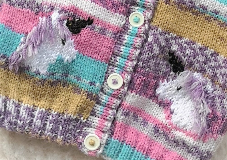 May include: Close-up of a knitted cardigan with a unicorn design. The sweater features a striped pattern in shades of pink, purple, blue, and tan. Two white unicorns with purple manes are embroidered on the front. The cardigan has three white buttons.
