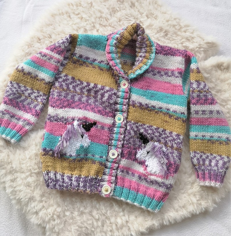 May include: A striped cardigan with a button closure and unicorn appliques. The cardigan is made in a variety of colors, including pink, purple, teal, and gold.