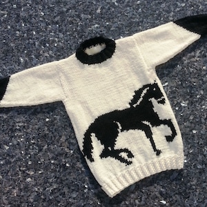 May include: A long-sleeved knit sweater in cream with a black horse silhouette on the front. The garment features a black ribbed crew neckline and black ribbed cuffs at the end of each sleeve, with a ribbed hem at the bottom of the sweater.
