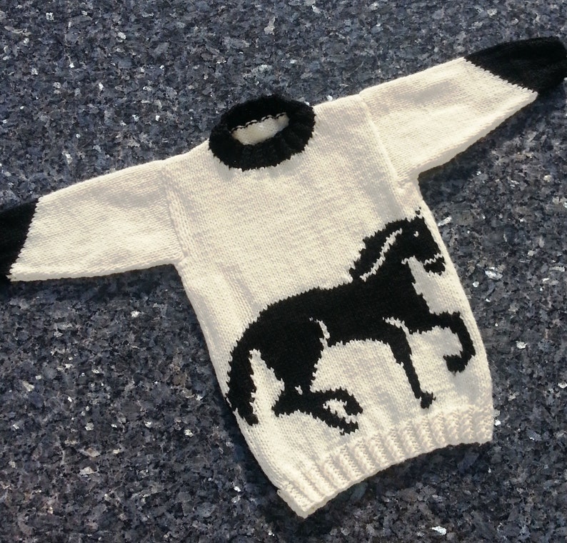 May include: A long-sleeved knit sweater in cream with a black horse silhouette on the front. The garment features a black ribbed crew neckline and black ribbed cuffs at the end of each sleeve, with a ribbed hem at the bottom of the sweater.