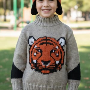 Op de afbeelding: Een kind draagt een gebreide beige trui met een tijgergezichtontwerp in oranje, zwart en wit. De trui heeft een hoge kraag en zwarte elleboogstukken. Het kind draagt ook een grijs en oranje gebreide muts.