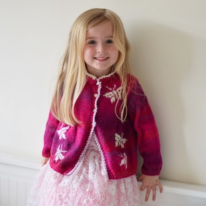 May include: A young girl wearing a pink cardigan with white butterfly embroidery. The cardigan has a button closure and is made of a soft, knitted fabric. The girl is wearing a white lace dress underneath the cardigan.