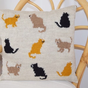 May include: Cream-colored knitted pillow featuring a pattern of various cat silhouettes in shades of gold, brown, and black. The cats are arranged in a repeating design across the pillow's surface. The pillow is square and appears to be made of a soft, textured yarn.
