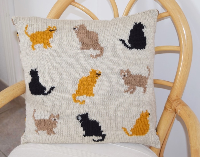 May include: Cream-colored knitted pillow featuring a pattern of various cat silhouettes in shades of gold, brown, and black. The cats are arranged in a repeating design across the pillow's surface. The pillow is square and appears to be made of a soft, textured yarn.