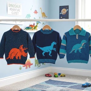 May include: Three long-sleeve sweaters hanging on wooden hangers. The left sweater is dark blue with an orange triceratops. The middle sweater is navy blue with a light blue dinosaur. The right sweater is blue with stripes and a dinosaur.