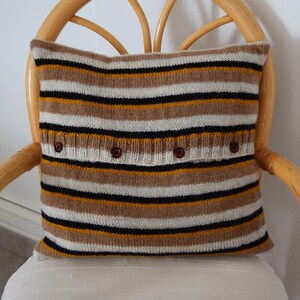 May include: A square pillow with horizontal stripes in shades of brown, black, white, and gold. The pillow has a knitted texture and is adorned with four dark red buttons. The pillow is placed on a white chair.