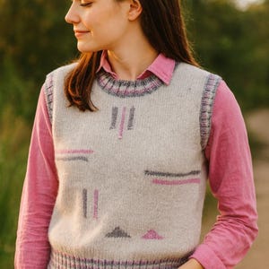 May include: A light gray knitted vest with geometric pink and gray accents. The vest has a ribbed hem and collar with a pink and gray pattern. Worn over a pink collared shirt and blue jeans.