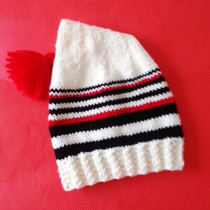 May include: A white knitted beanie featuring a ribbed cuff and horizontal stripes in black and red. The hat is topped with a bright red yarn pom-pom attached to the side, showcasing a classic knit texture throughout the main body of the headwear.