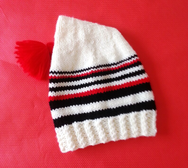 May include: A white knitted beanie featuring a ribbed cuff and horizontal stripes in black and red. The hat is topped with a bright red yarn pom-pom attached to the side, showcasing a classic knit texture throughout the main body of the headwear.